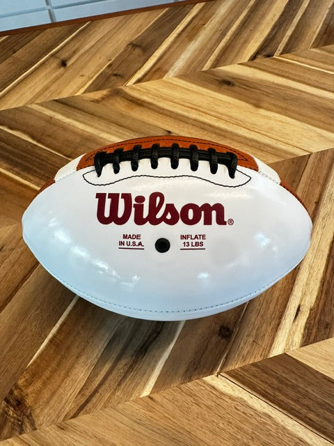 A white Wilson football with black laces and a brown top panel, resting on a wooden parquet floor.