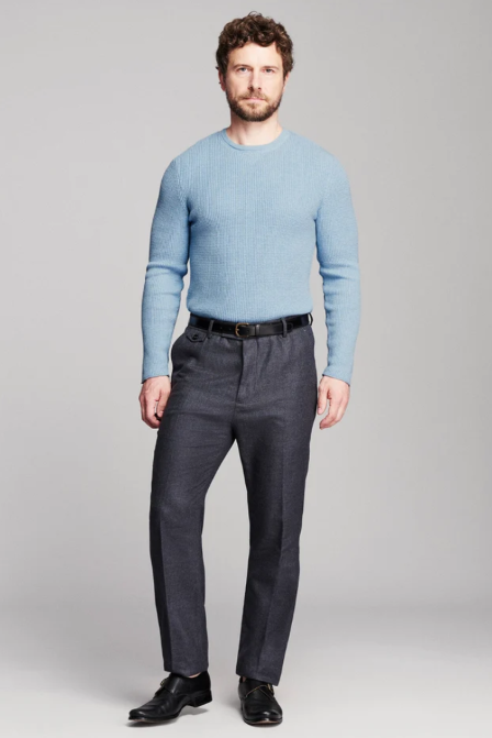 A man modeling a light blue crewneck knit sweater paired with dark gray trousers, a black belt, and black dress shoes, standing against a neutral gray studio background.