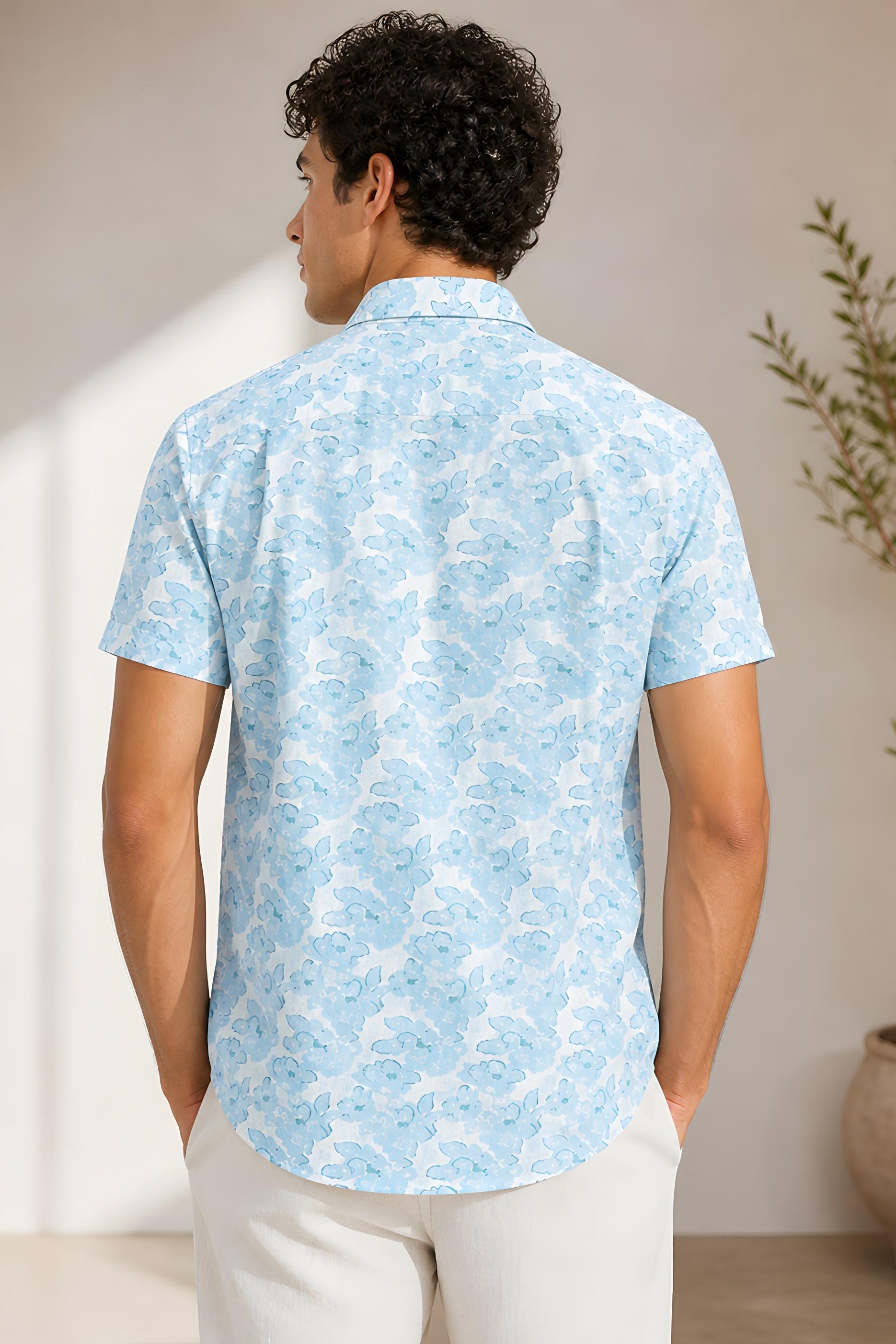 Man wearing a light blue patterned shirt standing in a room with a plant.