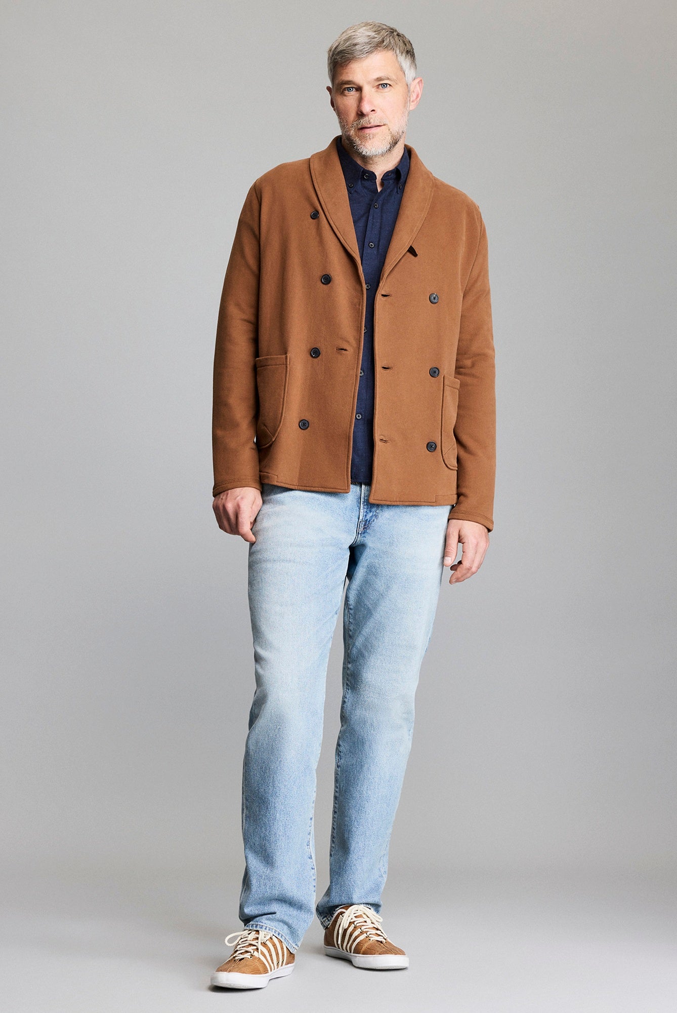 Man wearing a brown coat, blue shirt, and light blue jeans on a gray background