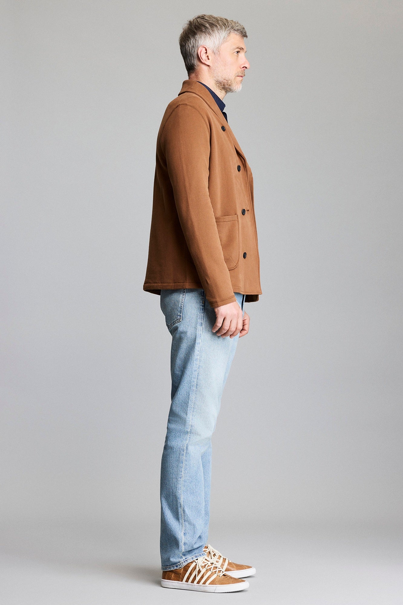 Man wearing a brown coat and light blue jeans standing against a plain background