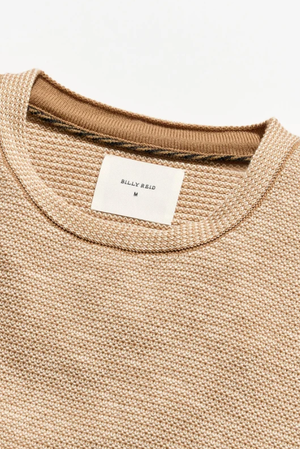 Close-up of a beige knit crewneck sweater with a white neck label reading BILLY REID M, set against a clean white background.