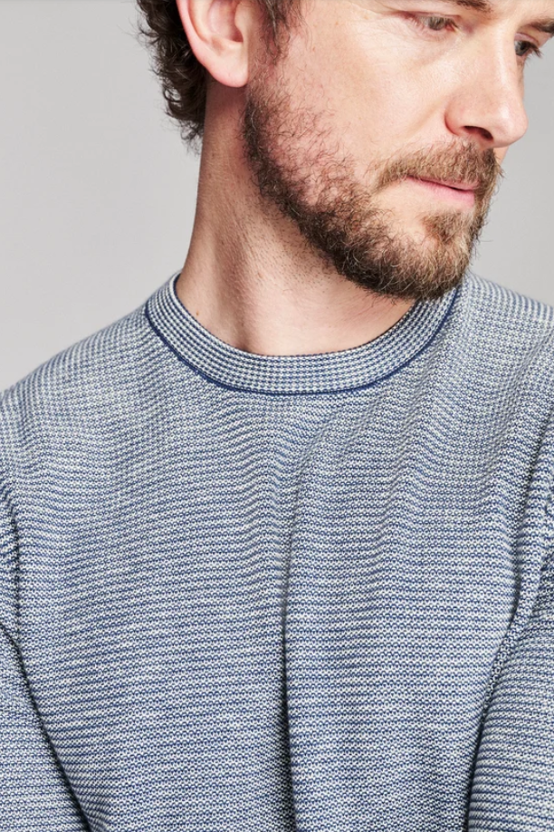 Close-up of a man wearing a textured knit crewneck sweater in a heathered blue and white pattern, set against a plain light gray background.