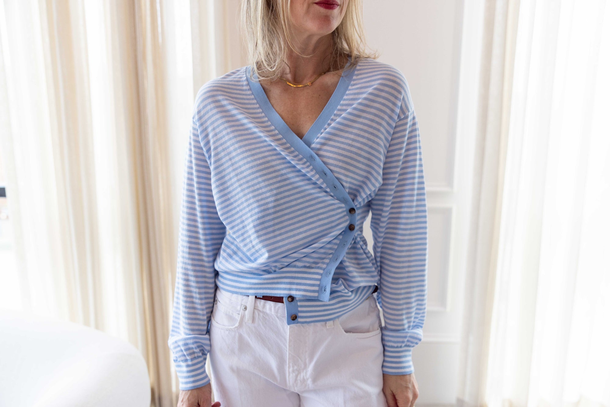 Close up of a woman wearing the ciao lucia jorgie cardigan standing in a room with white curtains.