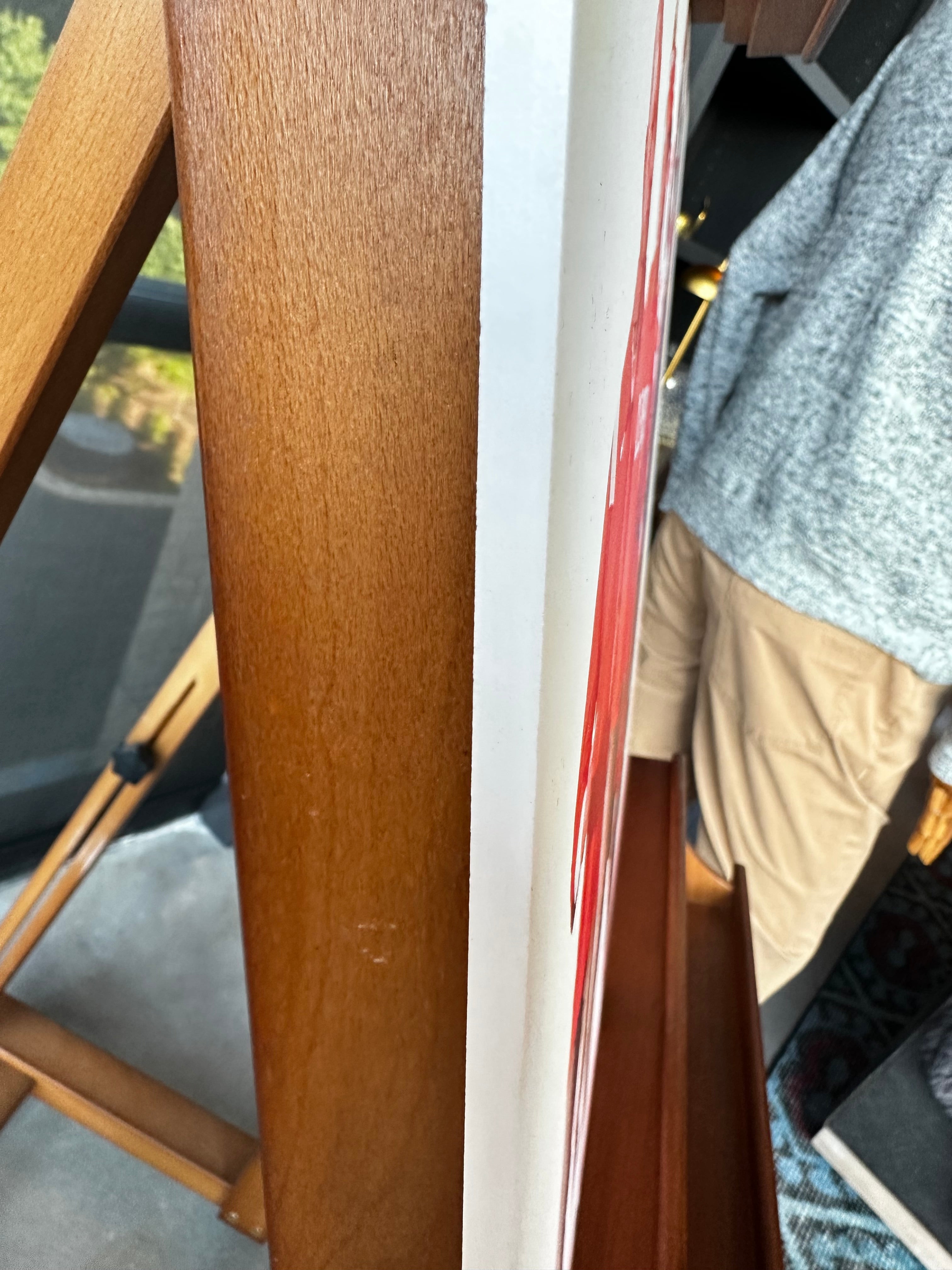 Close-up of a wooden furniture frame or leg in a warm medium brown finish, with part of a person wearing a gray sweater and khaki pants visible on the right side.