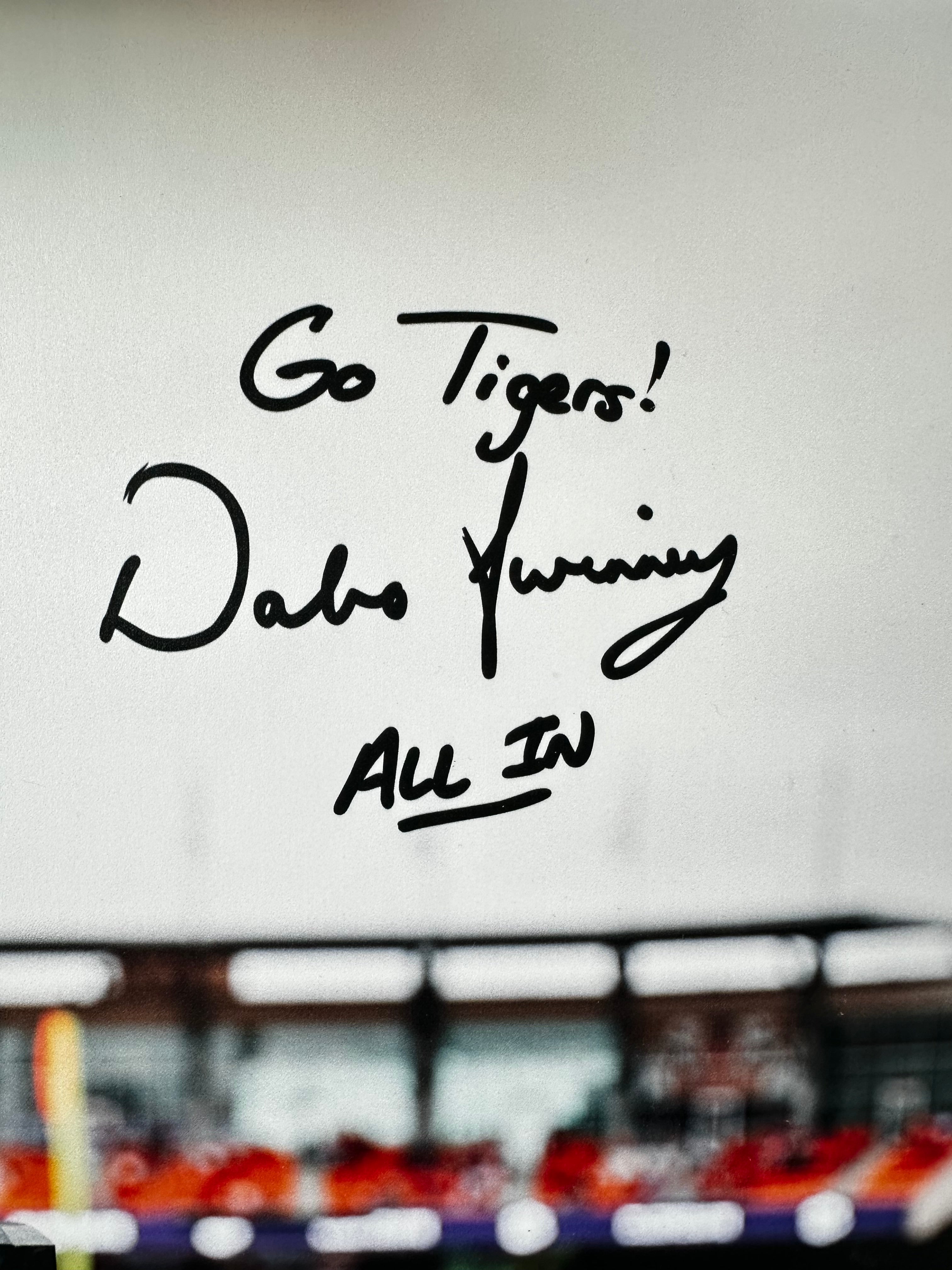 A close-up of a white surface with black handwritten text reading 'Go Tigers! Dabo Running ALL IN' over a blurred stadium background with red seating.