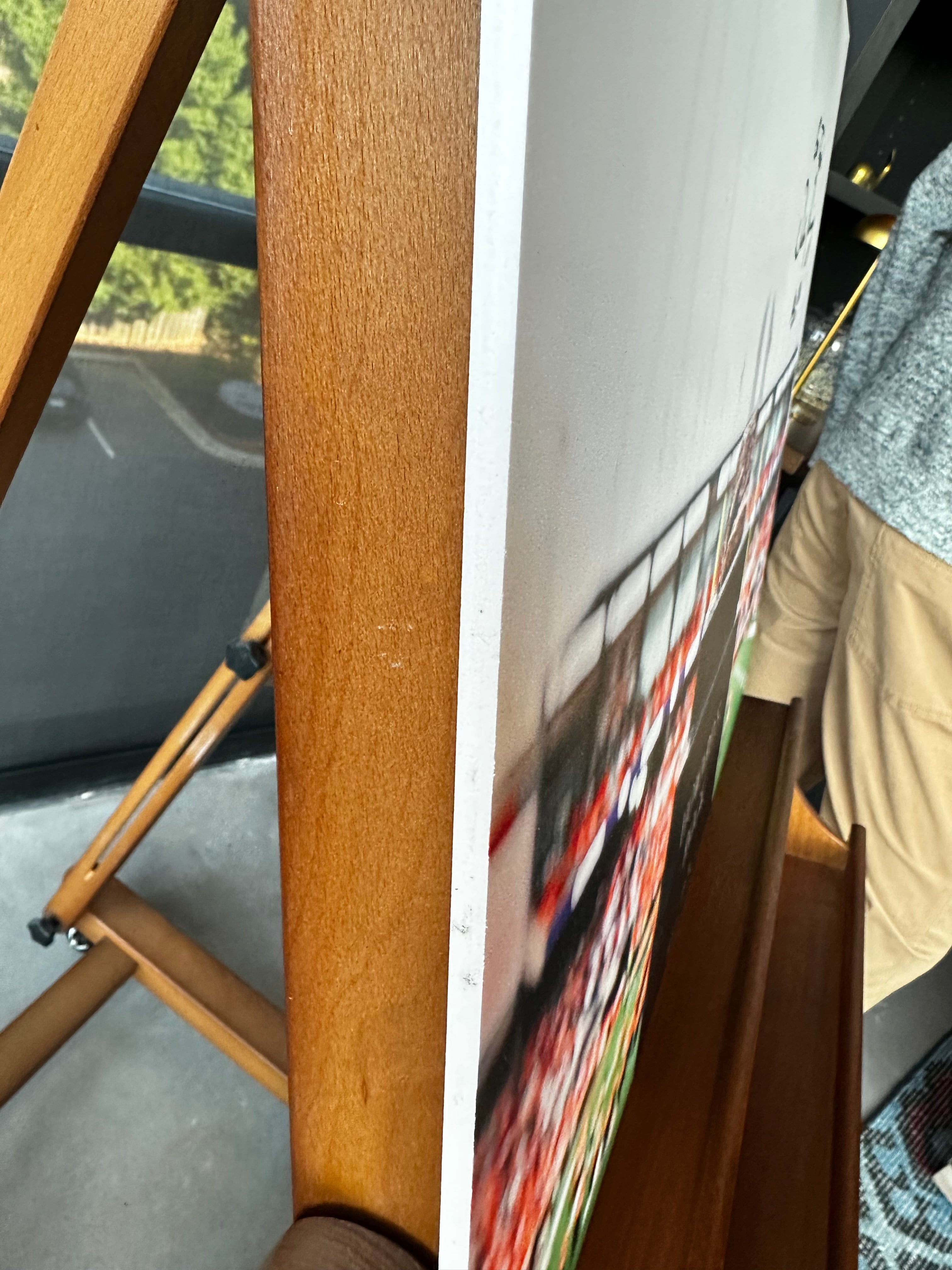 Close-up of a wooden easel holding a large vertical art print or canvas, with part of a person visible on the right side and an outdoor scene visible through a window on the left.