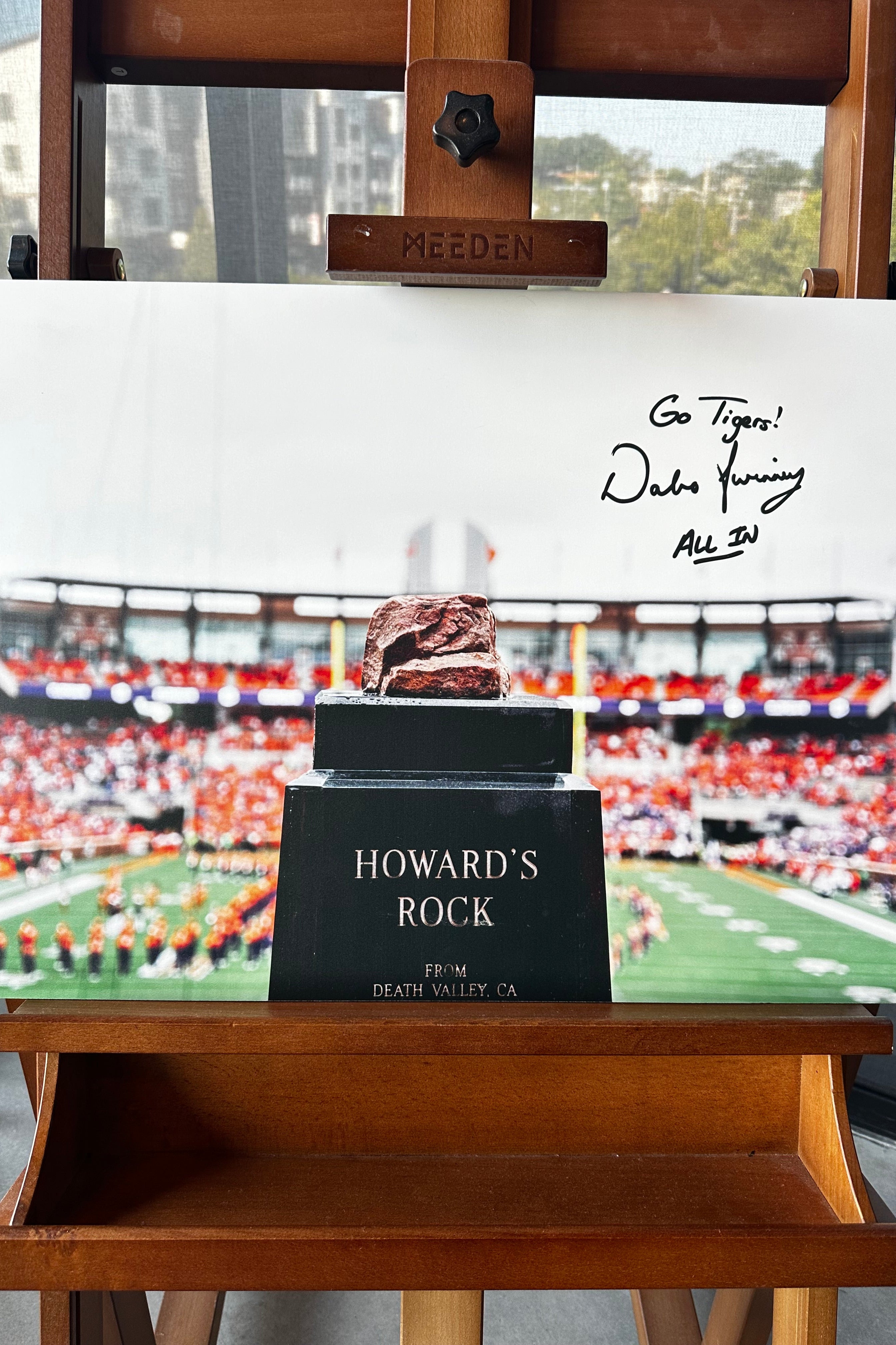 A large printed photo print displayed on a wooden Meeden easel. The print features a football stadium scene with a football player standing on a black pedestal labeled 'HOWARD'S ROCK', and handwritten black marker text reading 'Go Tigers! Deka Fanning All 34'.