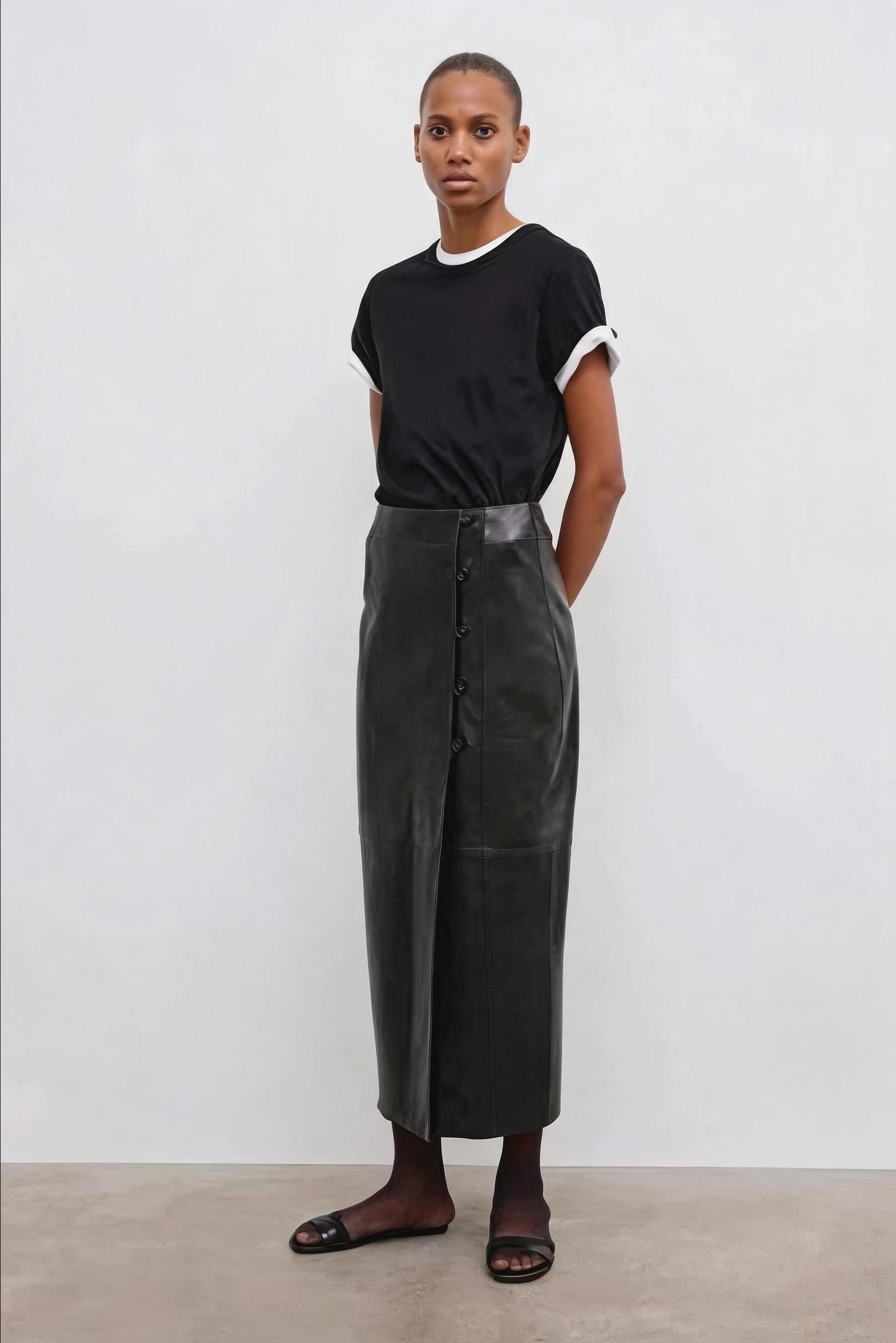 A model stands against a light gray studio wall wearing a black short-sleeve t-shirt with white ribbed trim and a long black leather-like midi skirt with front buttons and a wide black belt. She is wearing black slide sandals.