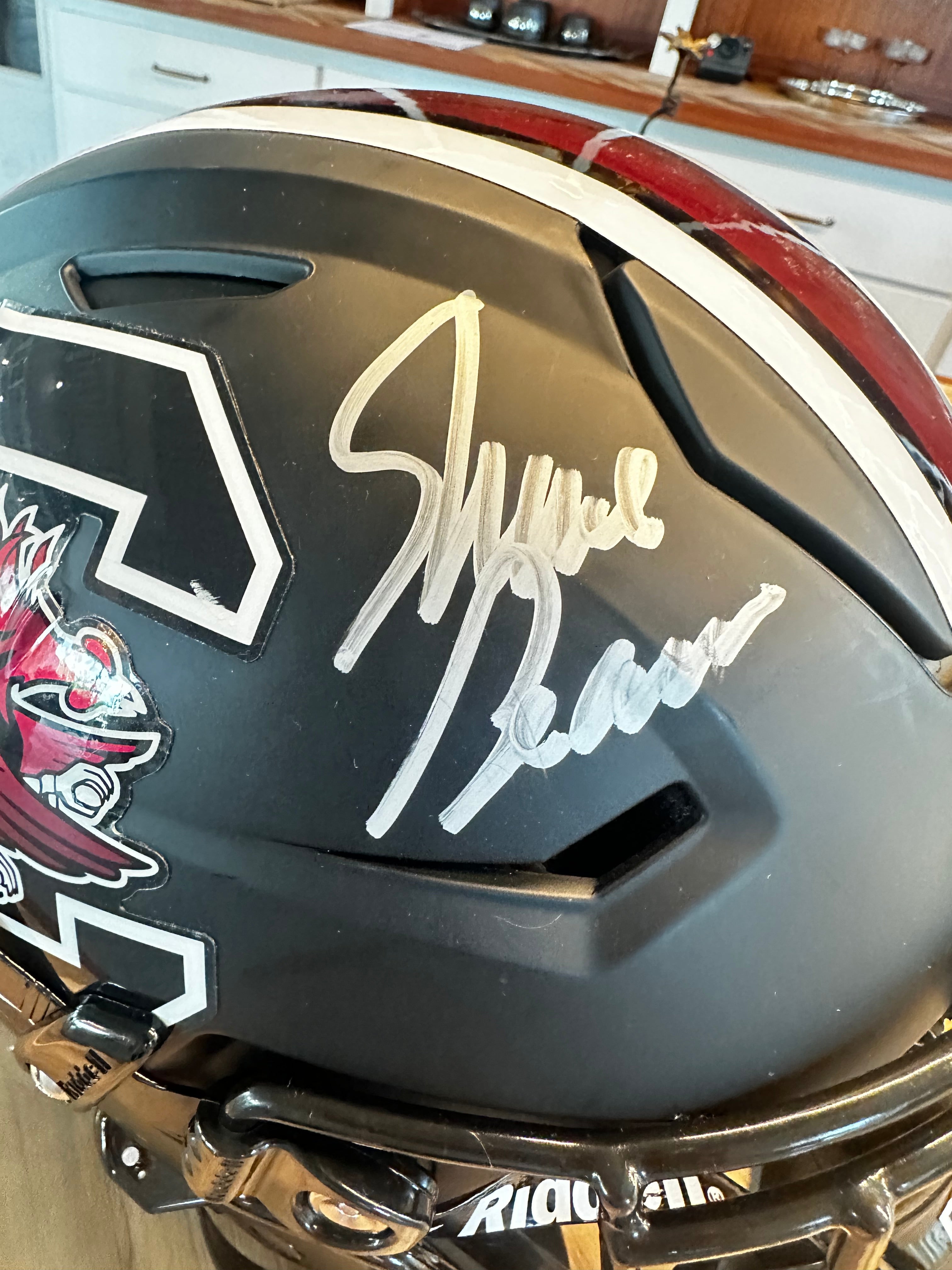 A close-up of a signed matte black football helmet with a white stripe, featuring a silver autograph on the side and part of the South Carolina gamecock logo visible.