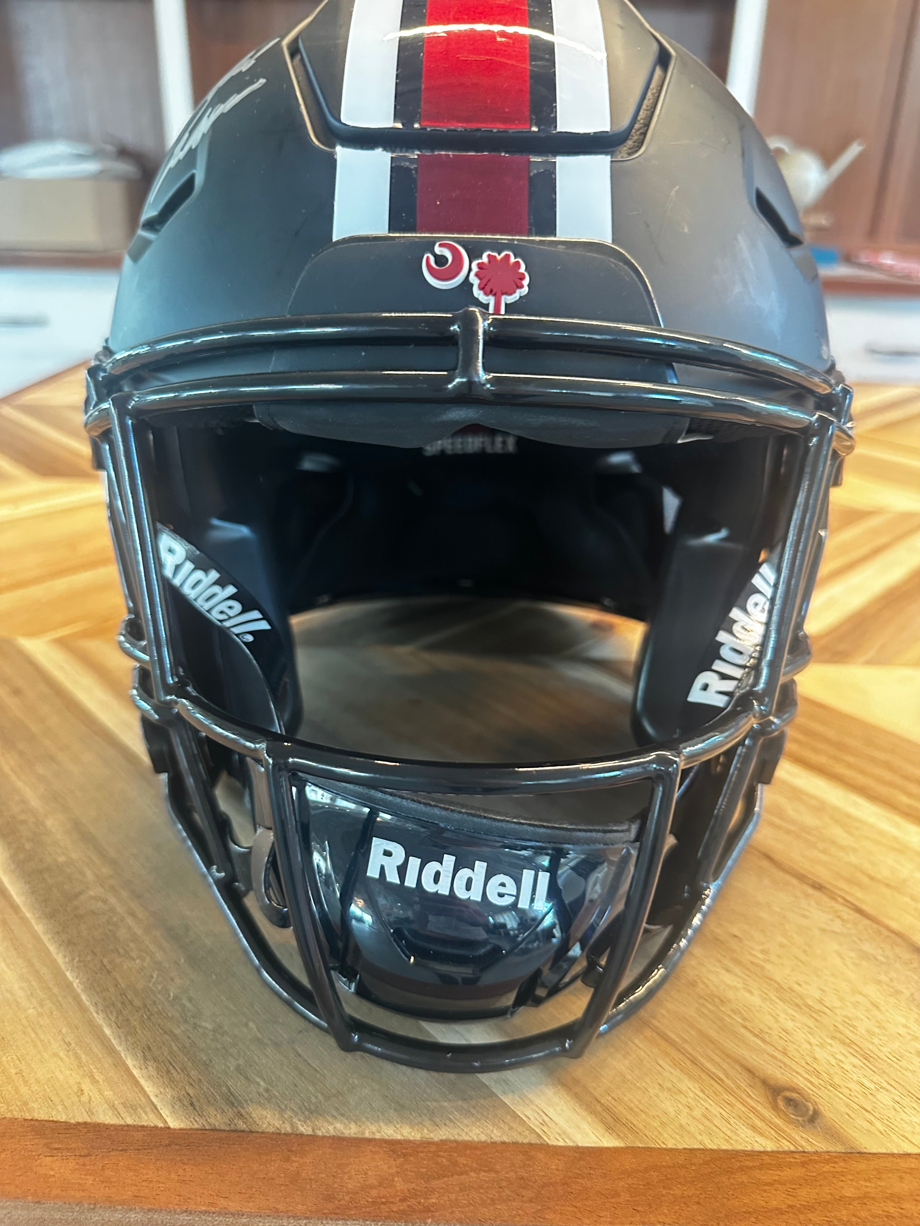 A black Riddell football helmet with a faceguard, featuring a red and white vertical stripe on top and a red maple leaf logo on the forehead, resting on a wooden table.