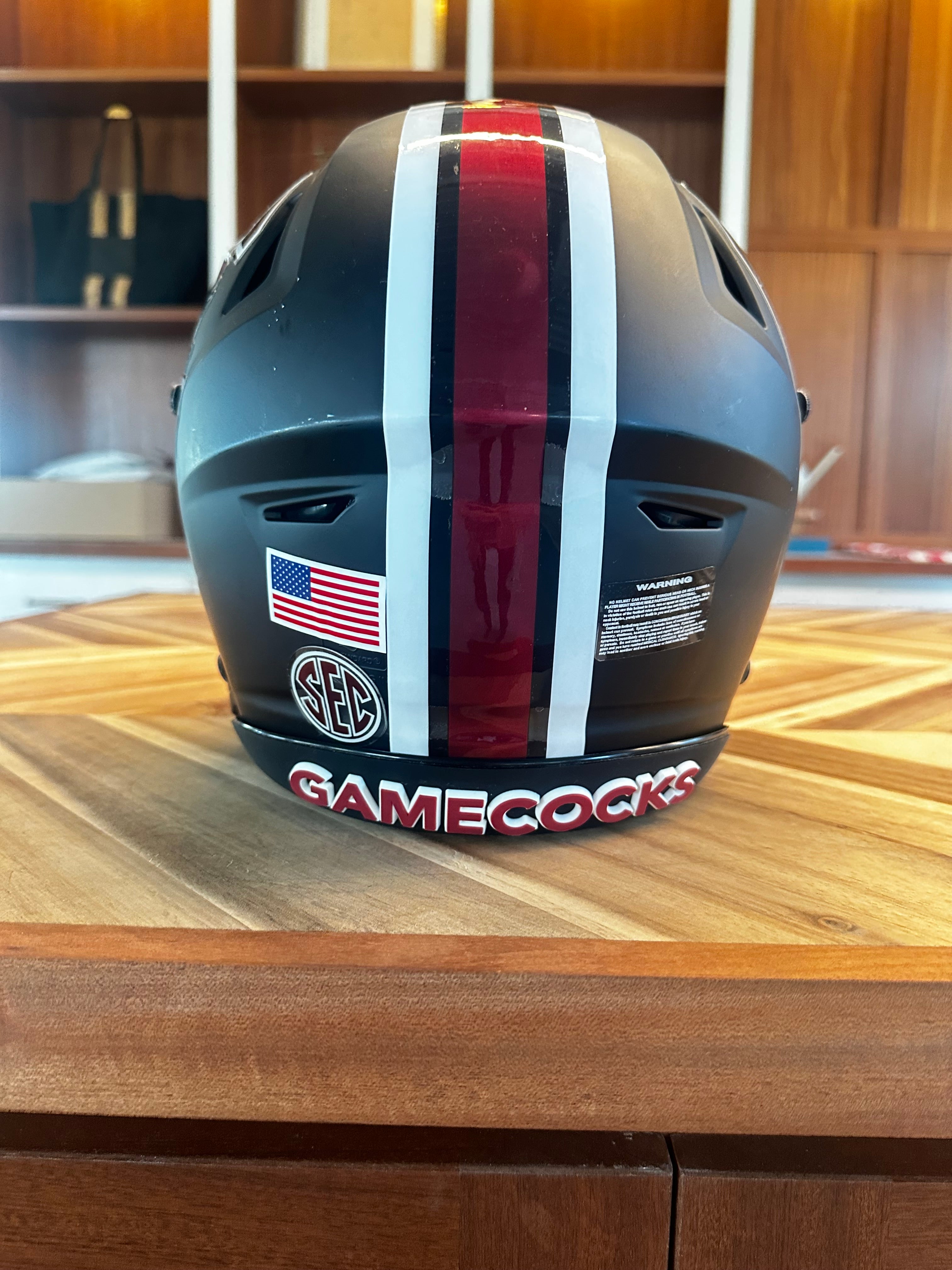 A black full-face helmet with a vertical red and white stripe, an SEC logo, an American flag decal, and 'GAMECOCKS' written in red on the lower edge, displayed on a wooden table.