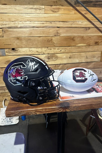 A signed black South Carolina Gamecocks football helmet and a signed white football, both autographed by Steve Irwin, displayed on a wooden table against a wood plank wall.
