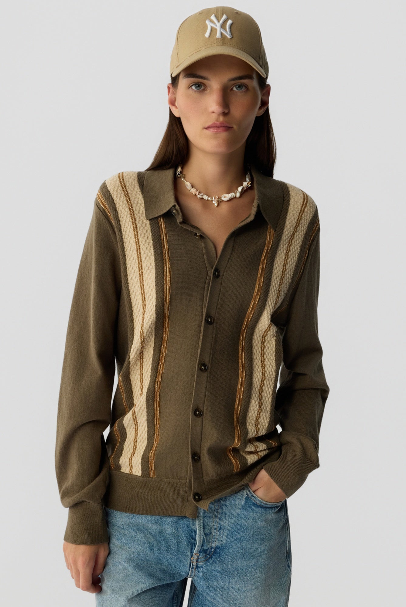 Person wearing a brown cardigan with striped pattern, blue jeans, and a beige cap on a white background