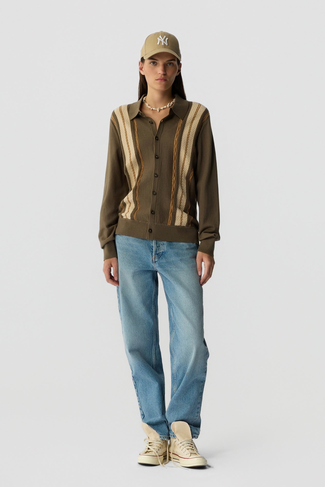 Person wearing a brown patterned shirt, blue jeans, and a cap on a white background