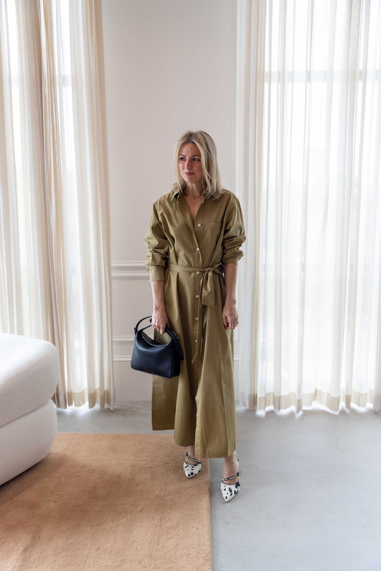 Woman in a green house of dagmar belted shirt dress standing in a room with white curtains and a white sofa.