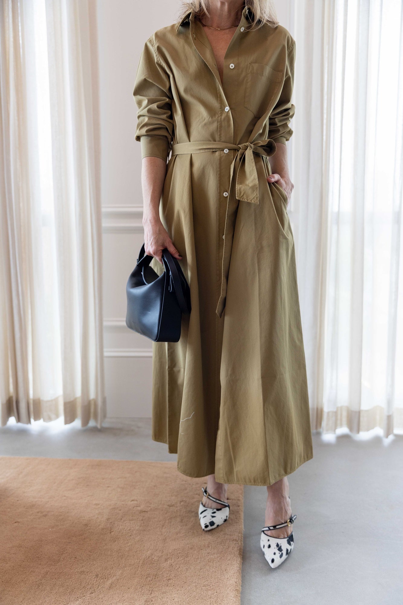 Close up of a woman wearing the green house of dagmar belted shirt dress standing in a room with a tan area rug and white curtains.