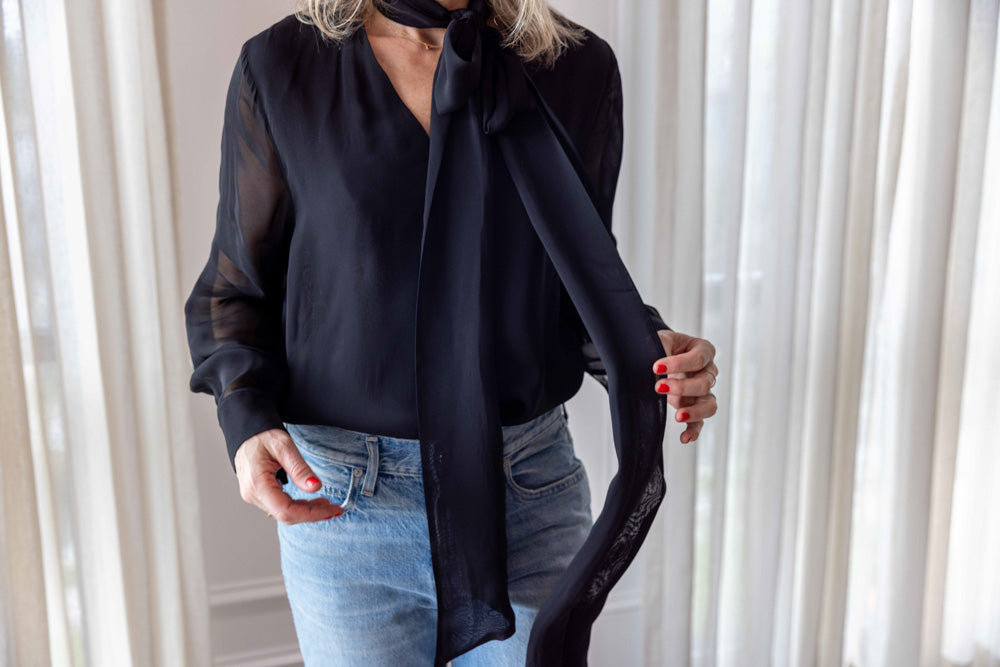 Dagmar Chiffon Scarf Blouse in black viscose, worn front view holding the long scarf tie, with light-wash denim