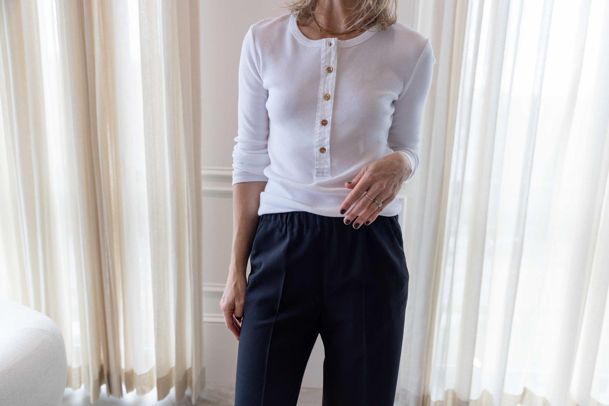 Close up of a woman wearing a house of dagmar henley top in white and house of dagmar elastic trouser standing in a room with white curtains and a white sofa.