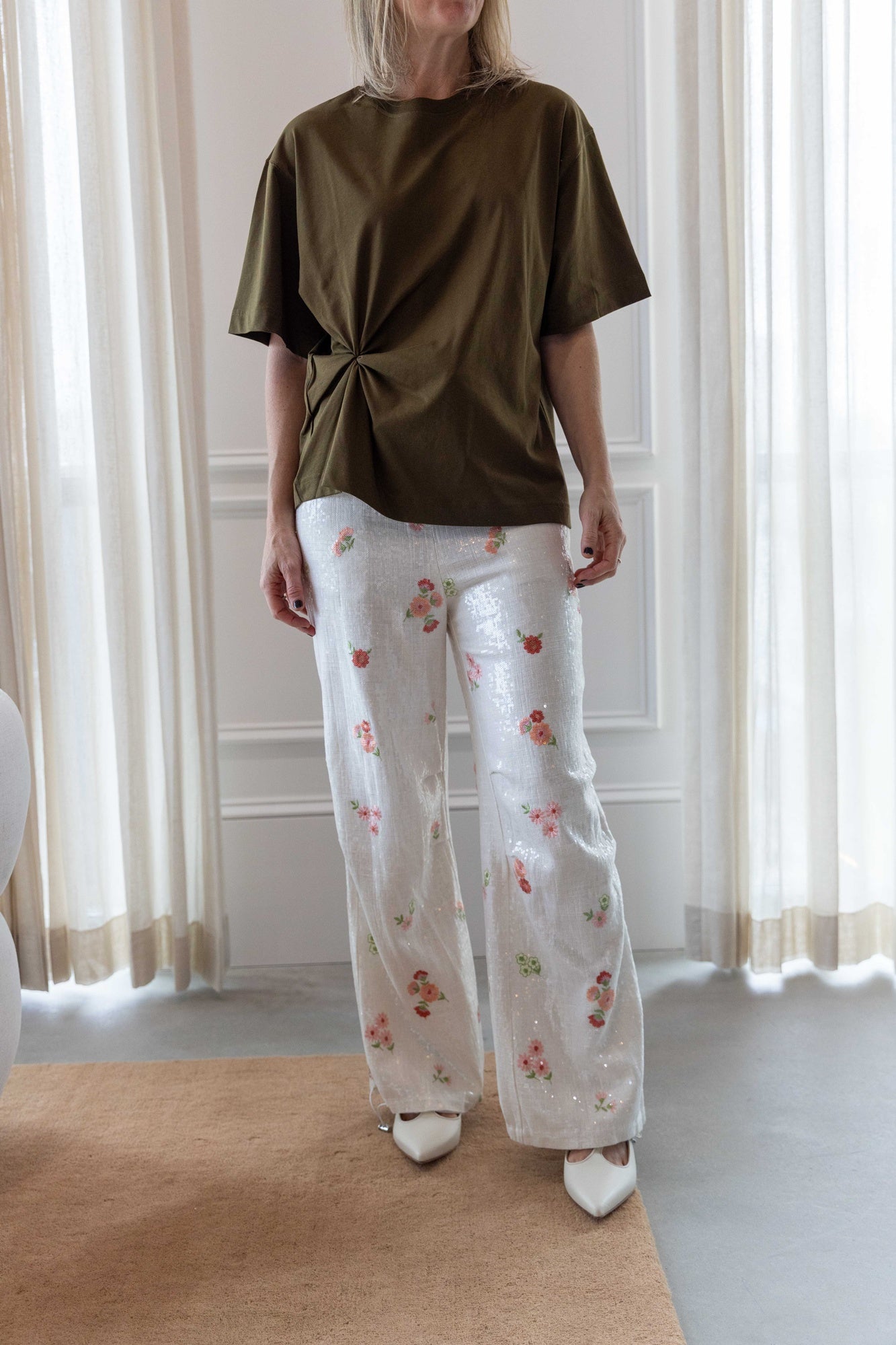 Woman wearing the house of dagmar knot t-shirt in green and the alfie grace sequin pant in floral standing in a room with white curtains and a tan area rug.