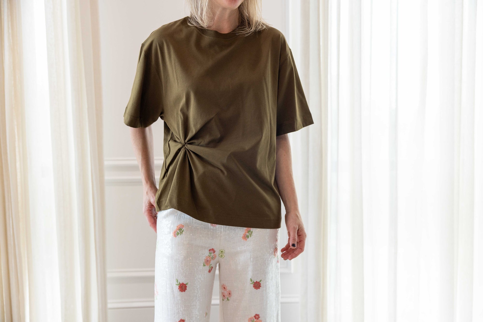 Close up of a woman wearing the house of dagmar knot t-shirt in green and the alfie grace sequin pant in floral standing in a room with white curtains.