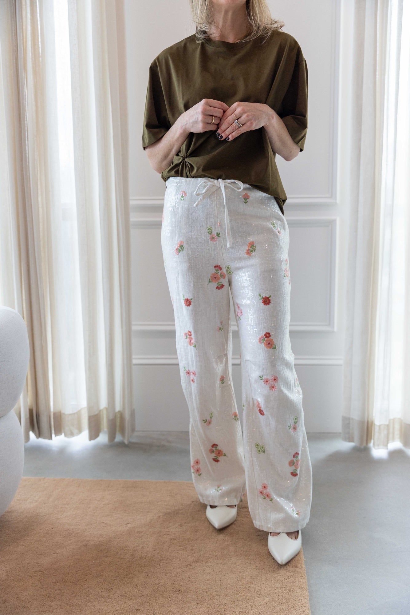 Woman wearing the house of dagmar knot t-shirt in green and the alfie grace sequin pant in floral standing in a room with white curtains and a tan area rug.