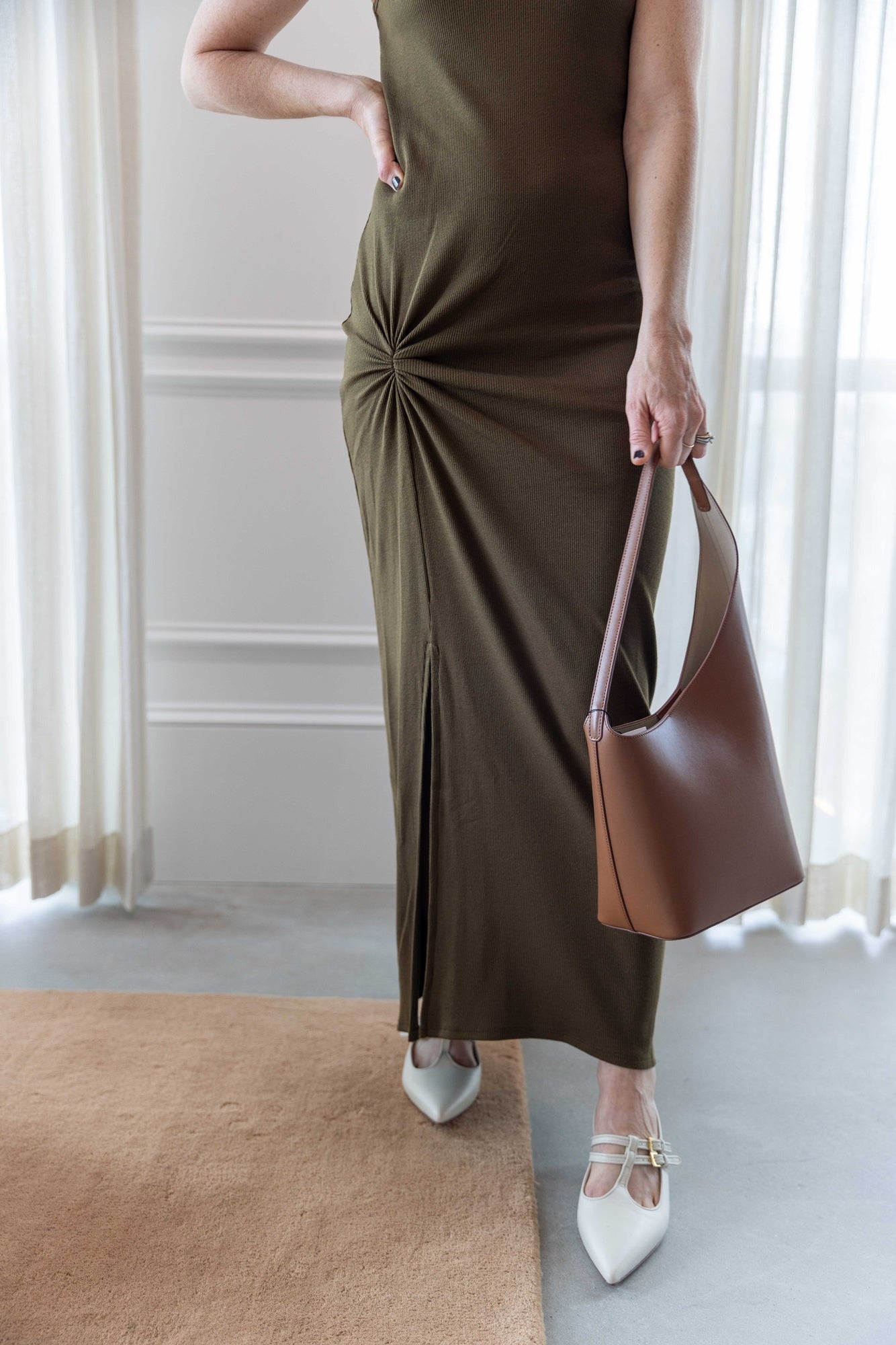 Close up of a woman wearing the house of dagmar knot tank dress and holding the aesther ekme new demi lune handbag house standing in a room with white curtains and a tan area rug.
