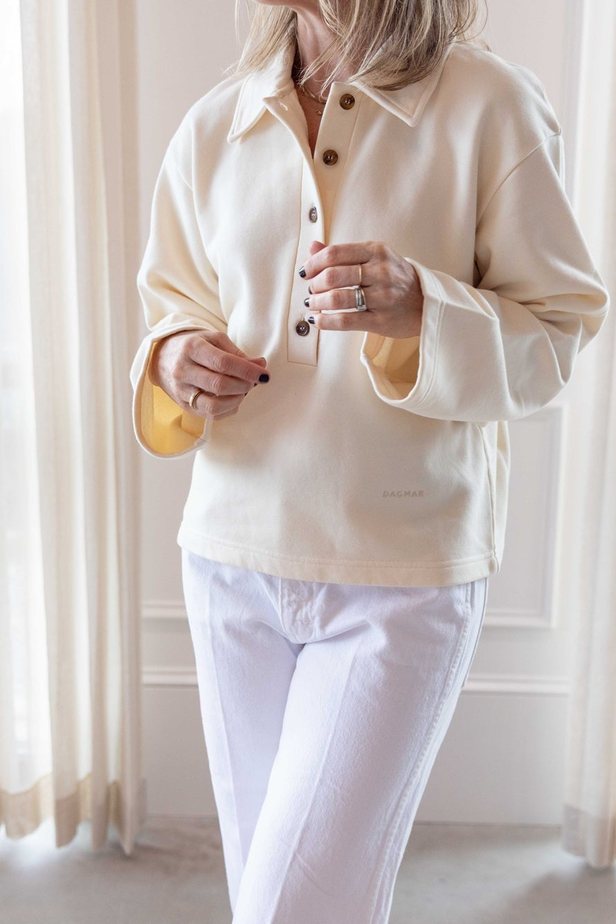 Close up image of a woman wearing the house of dagmar polo sweatshirt and amo denim mid-rise relaxed jean standing in a room with a white sofa and white curtains.