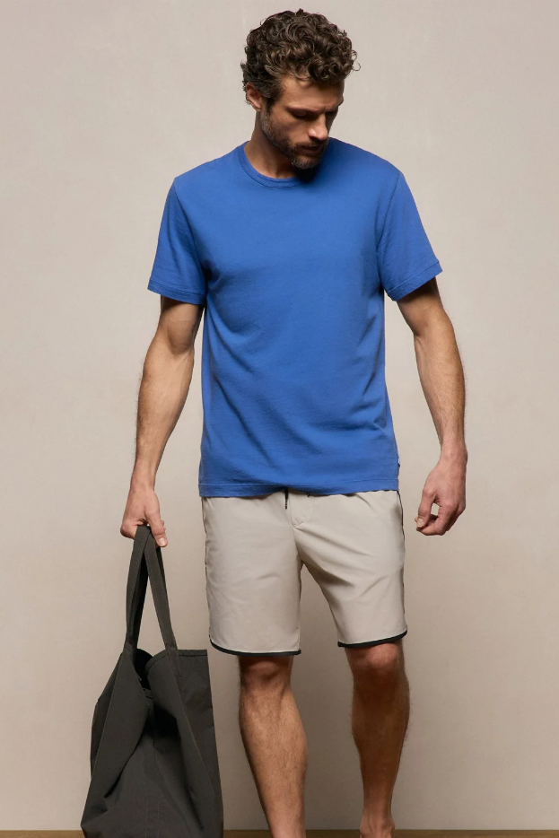 Man wearing a blue t-shirt and beige shorts holding a black bag against a plain background