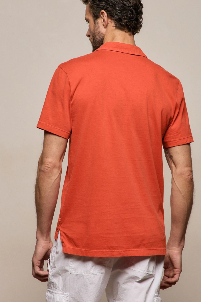 Man wearing an orange polo shirt and white pants against a beige background