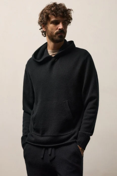 A man with curly brown hair and a beard wearing a black knit pullover hoodie and matching black sweatpants, standing against a plain light beige wall.