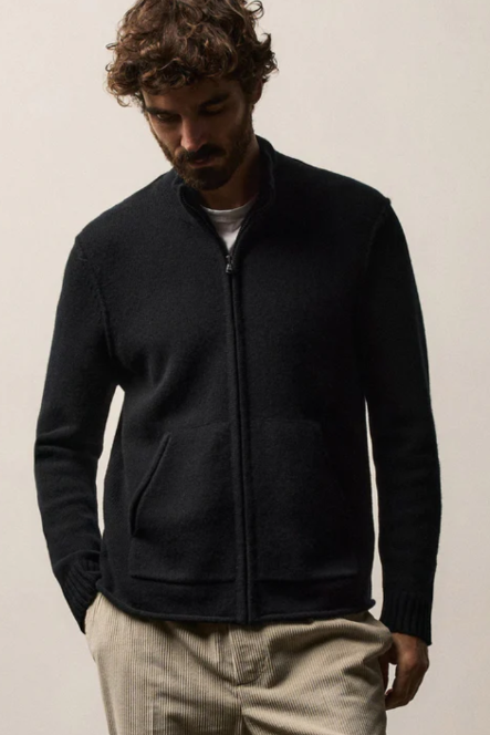 A man wearing a black zip-up knit sweater and beige trousers, standing against a plain light beige background.