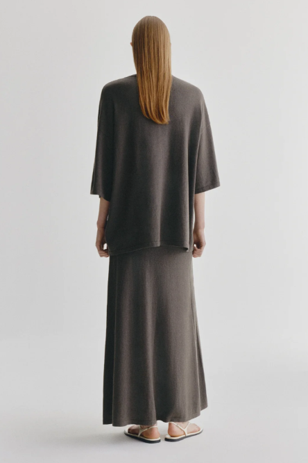 A model viewed from behind wearing a matching dark brown knit two-piece set consisting of a short-sleeve top and a long skirt, paired with neutral sandals, against a light gray studio background.