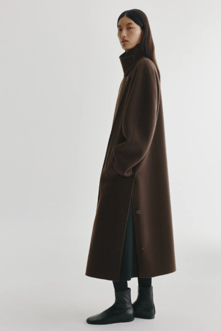 A model wearing a long, dark brown wool coat with a high collar, styled with black trousers and black leather boots against a plain light gray studio background.