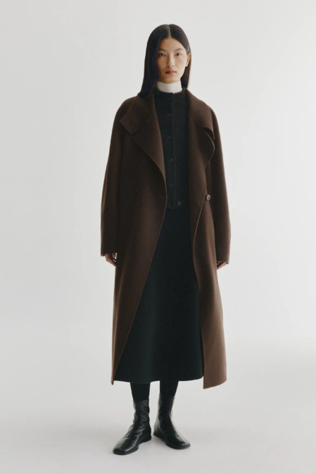 A model stands against a light gray studio background wearing a long brown overcoat over a black dress, black turtleneck, black socks, and black ankle boots.