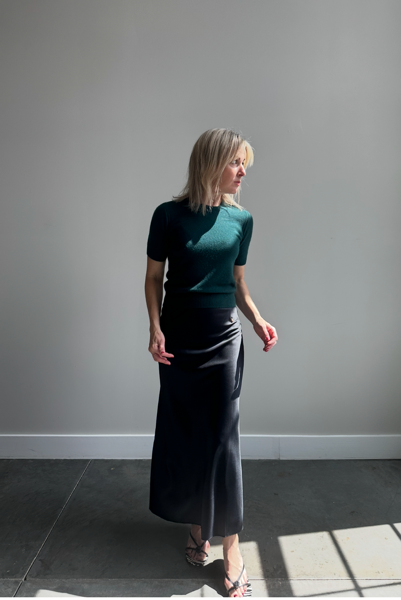 Woman wearing a green top and dark skirt standing against a plain wall.