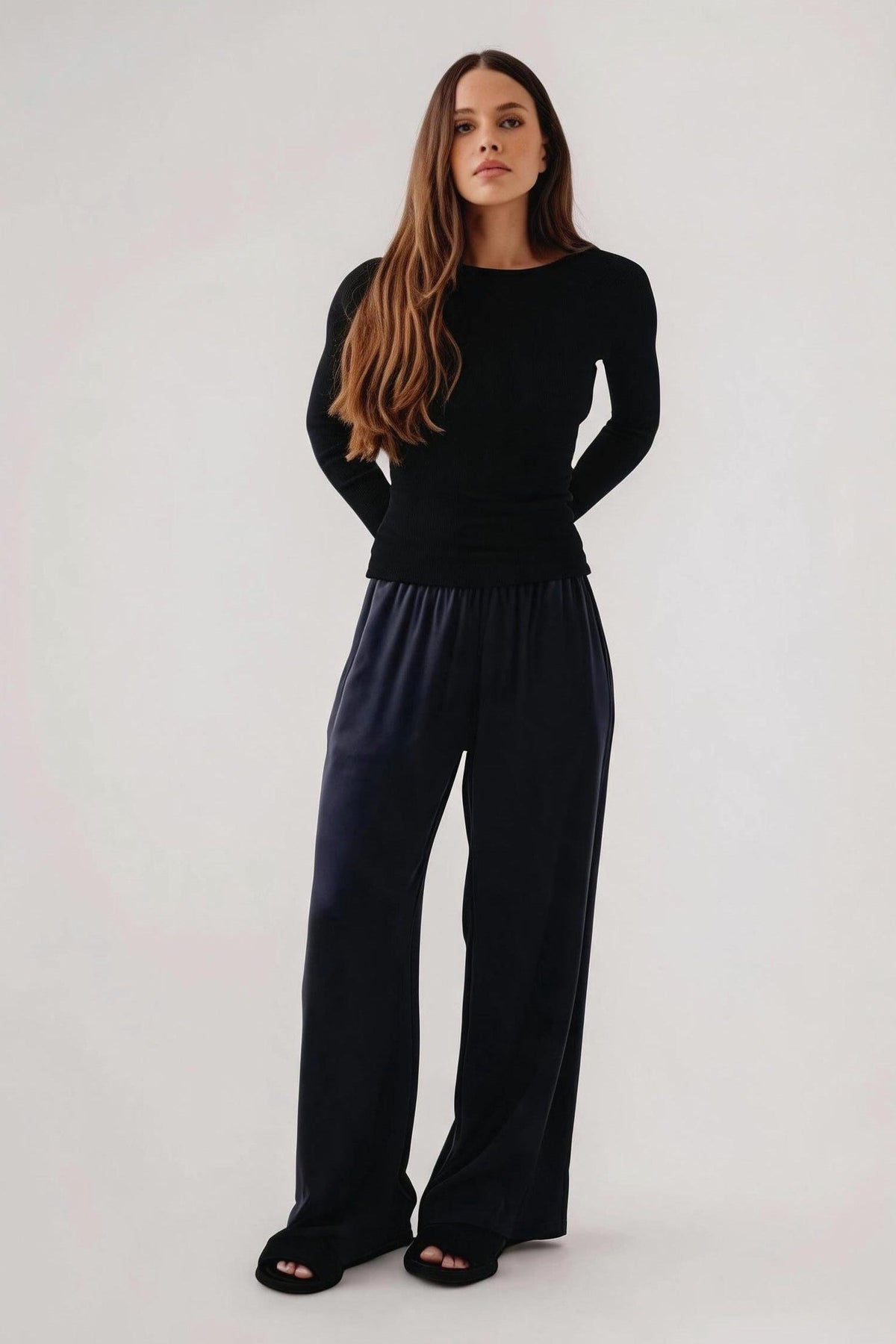 A woman modeling a black long-sleeve top and dark navy wide-leg pants against a plain light gray studio background.