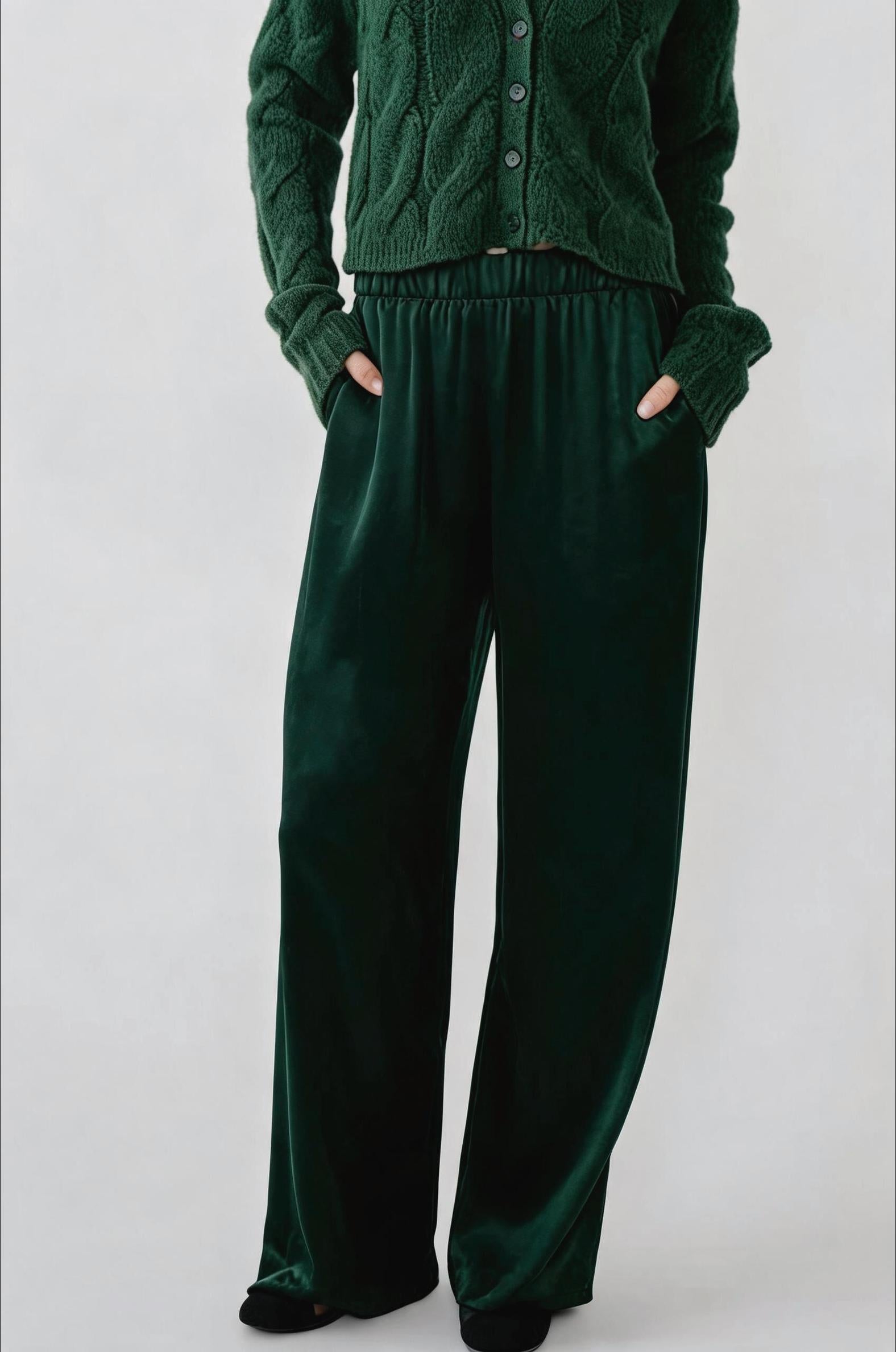 A person wearing a dark forest green knit cardigan and matching wide-leg velvet pants, standing against a light gray studio background with hands in the pants pockets.