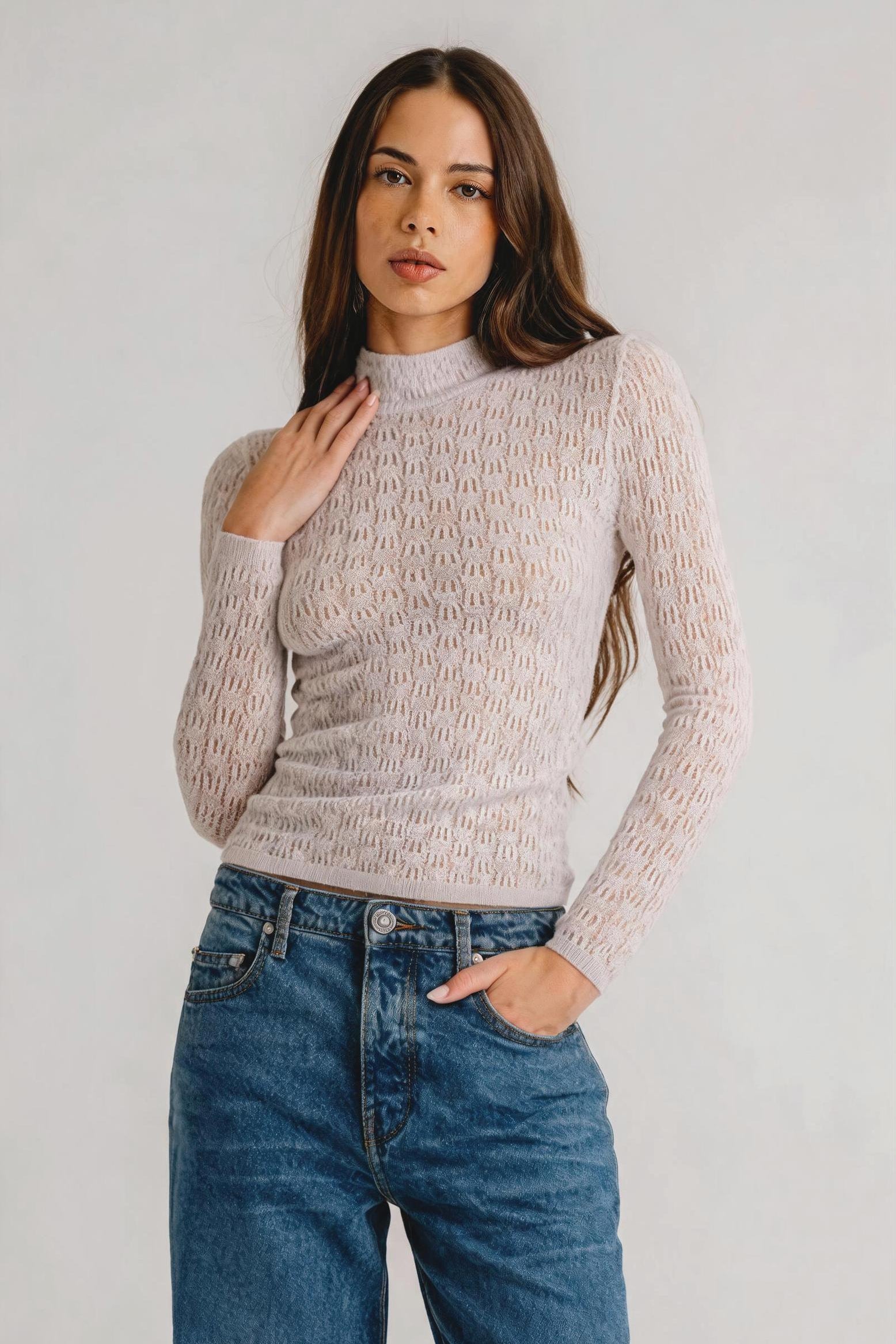 A woman modeling a pale pink textured knit mock-neck sweater with long sleeves, paired with blue jeans, against a light gray studio background.