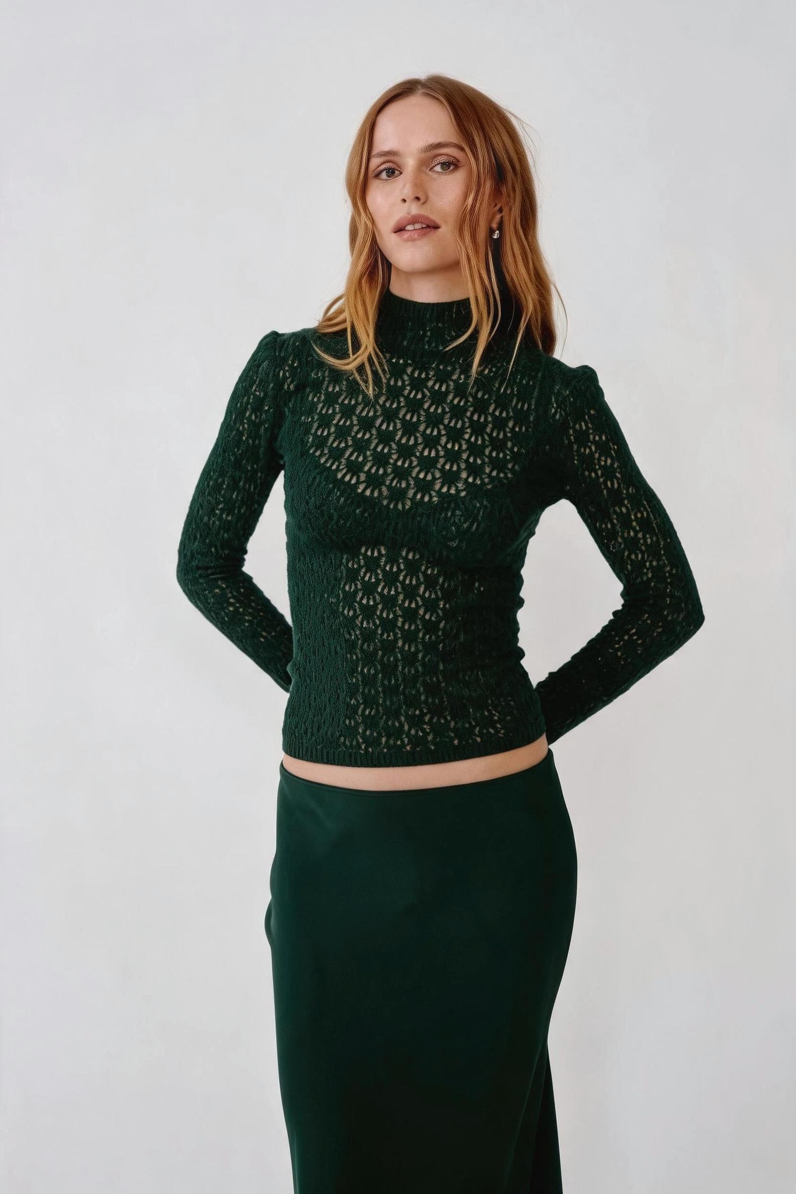 A woman modeling a dark green long-sleeve knit mock-neck top with a textured geometric pattern, paired with a matching dark green skirt, against a plain light gray studio background.