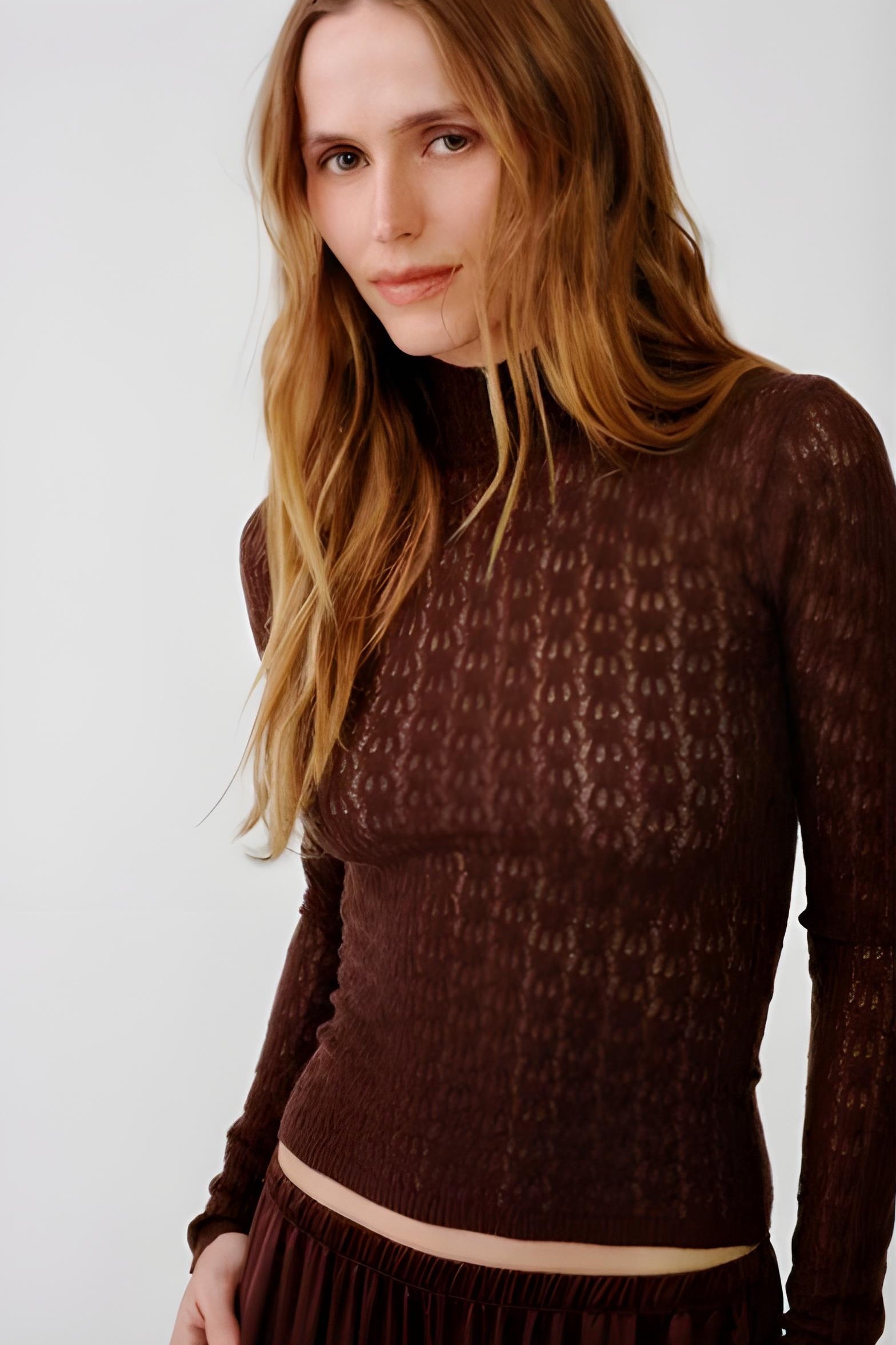 A woman wearing a dark brown textured knit mock neck long-sleeve top, shown from the chest up against a plain light gray background.