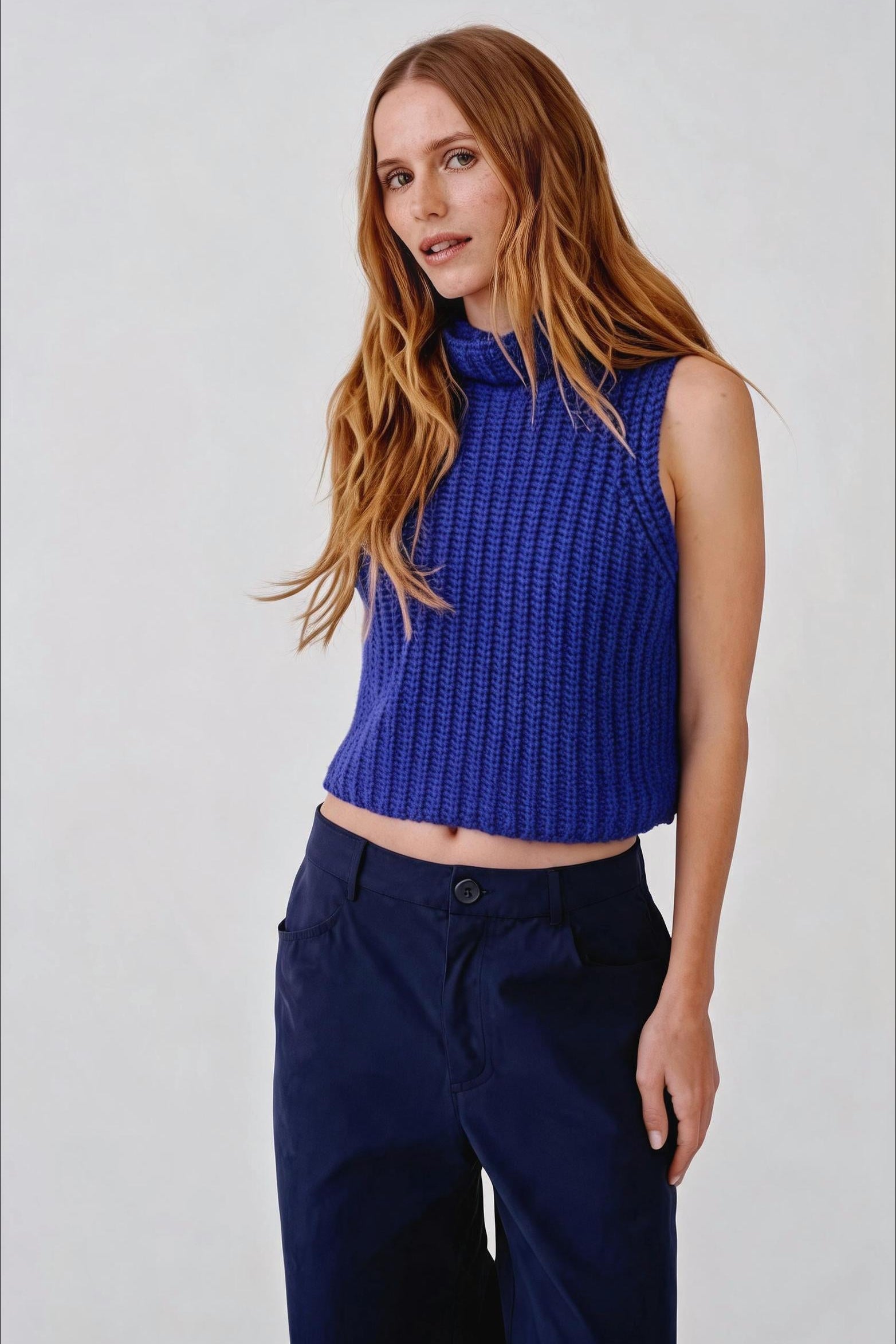 A woman with long light brown hair wearing a textured royal blue sleeveless mock-neck crop top and dark navy trousers, standing against a light gray studio background.