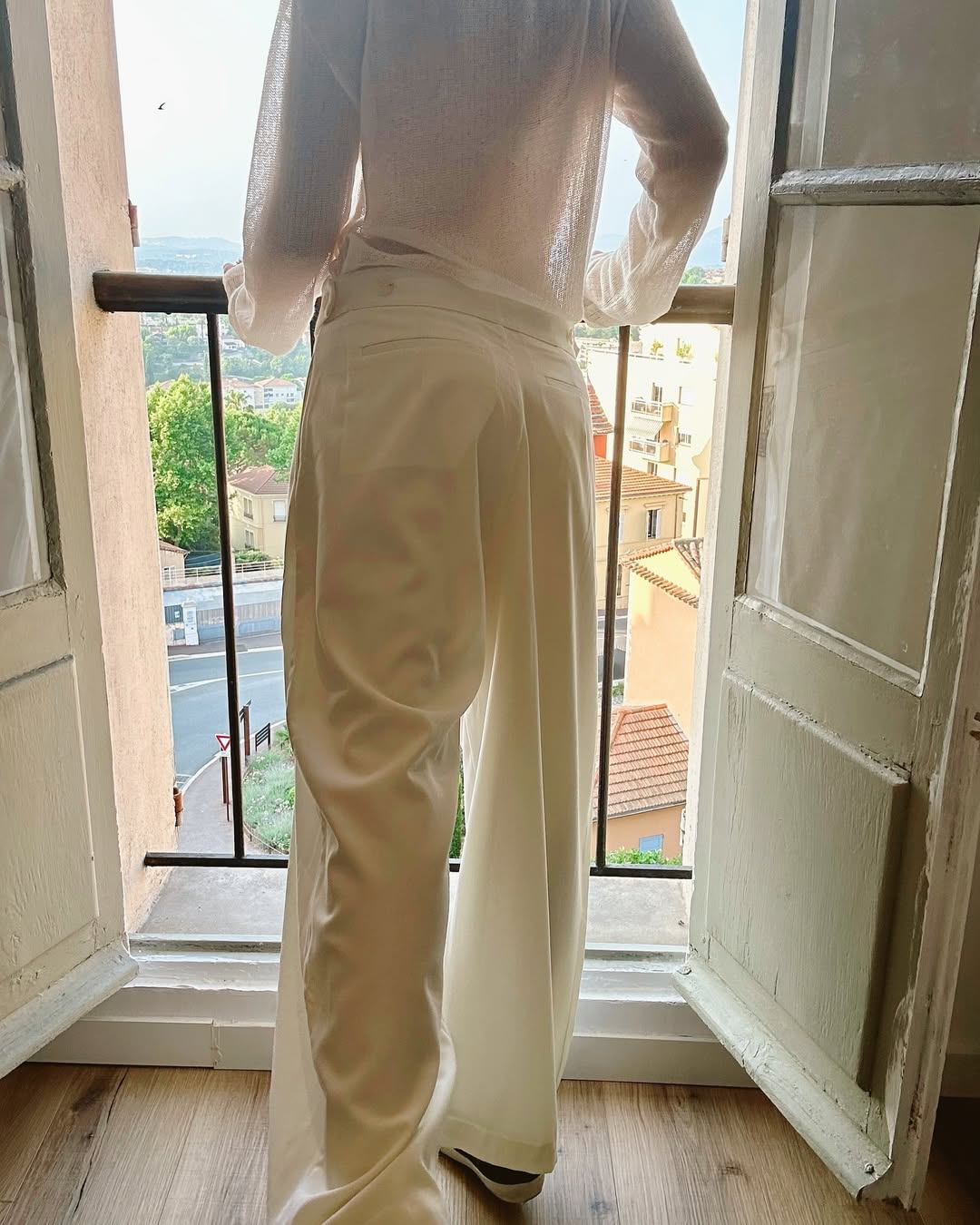 Person wearing a white outfit standing in a room with an open door and view of buildings outside.