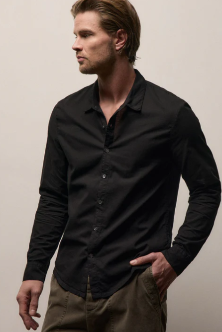 A man wearing a black long-sleeve button-down shirt and olive green pants, standing against a plain beige background.