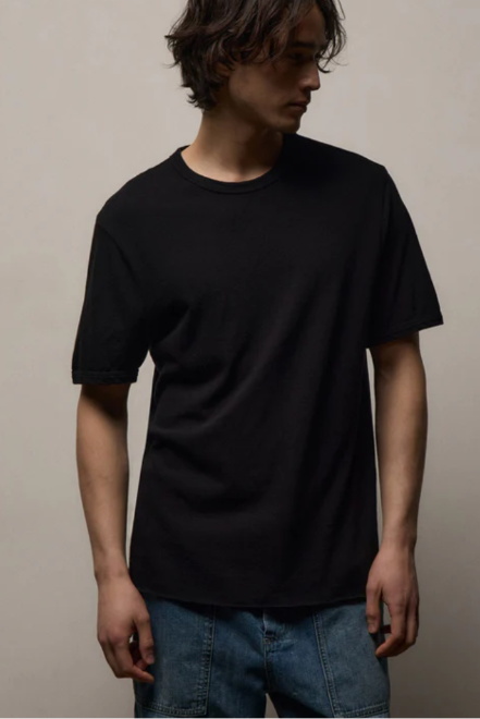A man wearing a plain black short-sleeve crewneck t-shirt and blue jeans, standing against a neutral beige background.