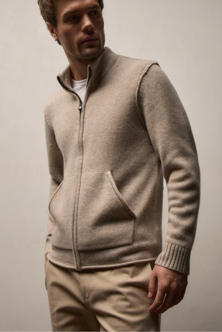 A man wearing a light beige zip-up knit sweater vest over a white t-shirt and beige trousers, photographed against a neutral light gray studio background.