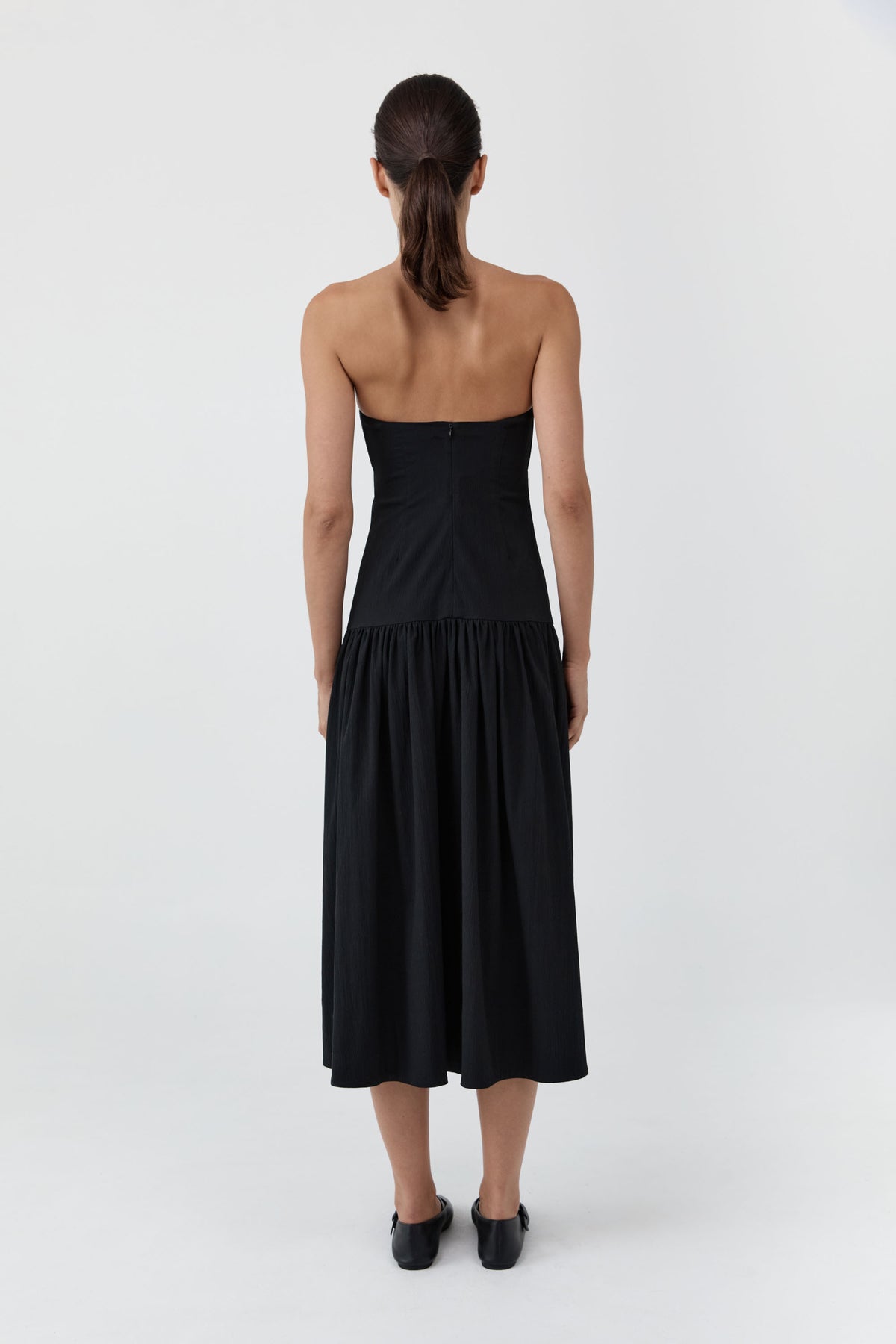 A woman viewed from the back wearing a strapless black midi dress with a gathered waist and black shoes, standing against a light gray studio background.