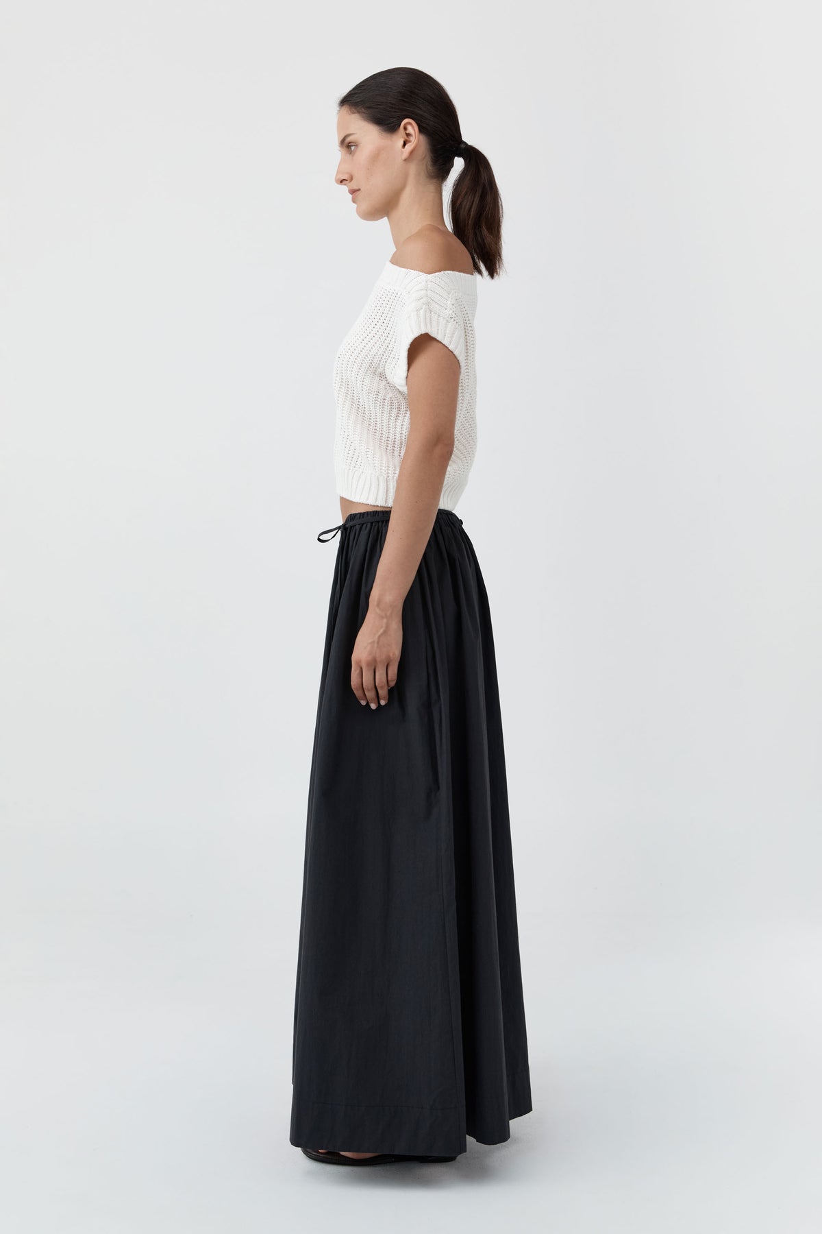 A woman in profile wearing a white ribbed short-sleeve top and a long black skirt, standing against a light gray studio background.