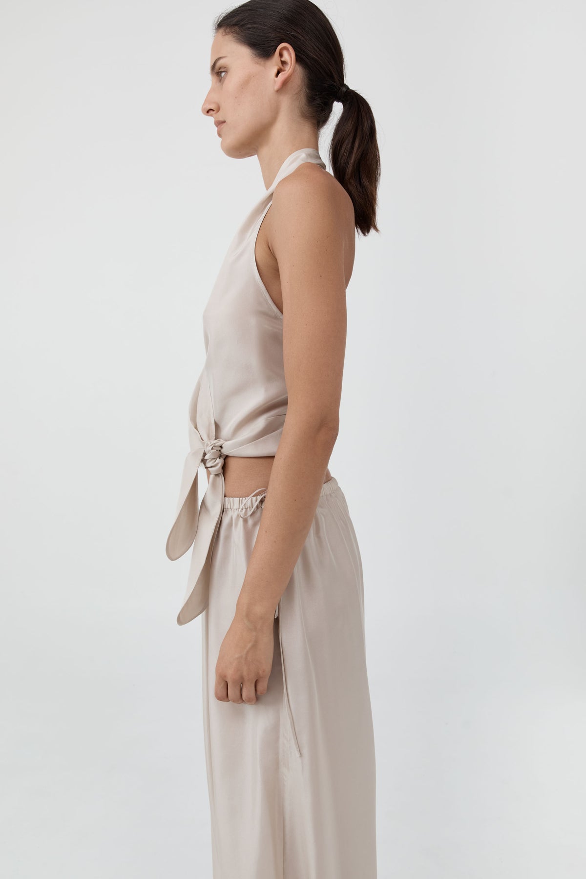 A woman shown in side profile wearing a light beige sleeveless wrap-style top and matching high-waisted trousers against a plain light gray studio background.