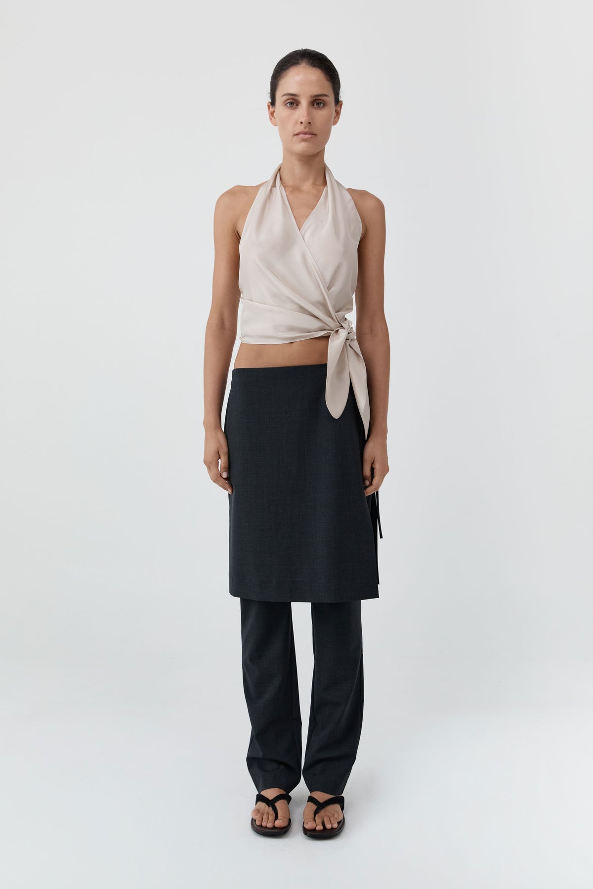 A model stands against a light gray studio background wearing a sleeveless cream halter wrap top tied at the waist, dark charcoal wrap skirt, matching dark pants, and black sandals.